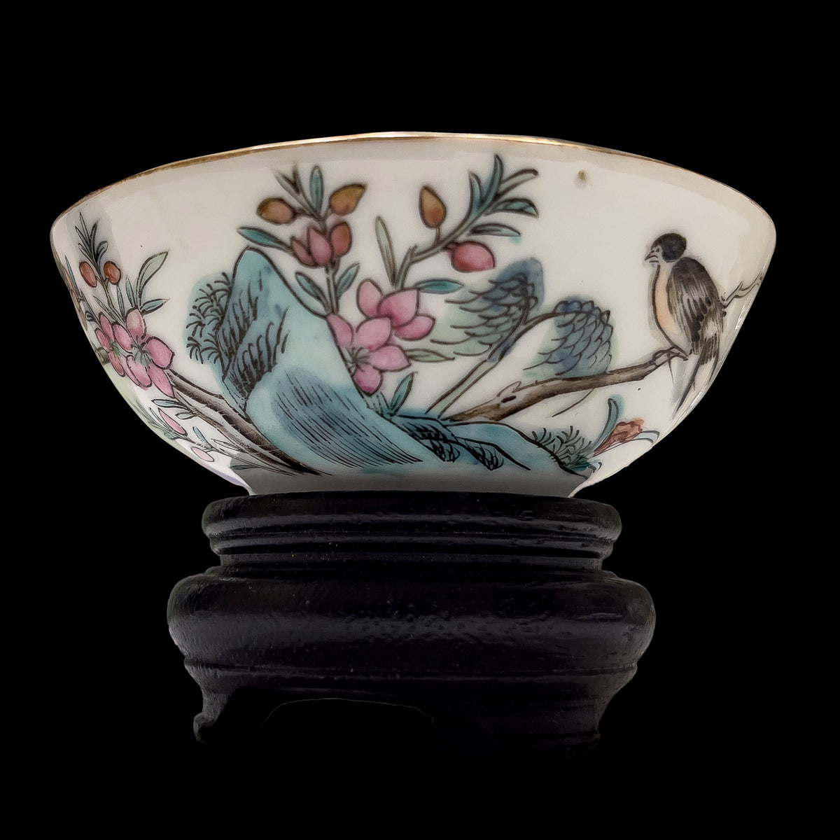 An antique Chinese famille rose porcelain wine teacup showcasing a hand-painted songbird on a plum branch with a gilded rim, resting on a carved dark wood stand over a black ground.