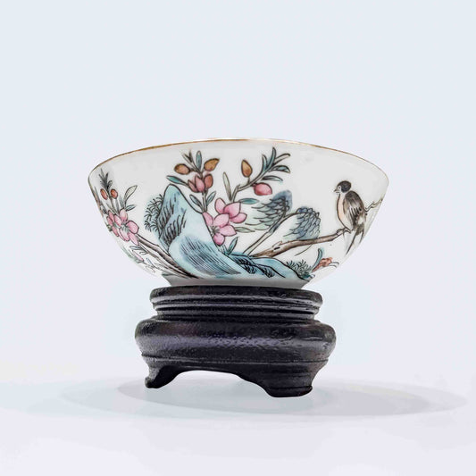 An antique Chinese famille rose porcelain wine teacup showcasing a hand-painted songbird on a plum branch with a gilded rim, resting on a carved dark wood stand over a neutral background.
