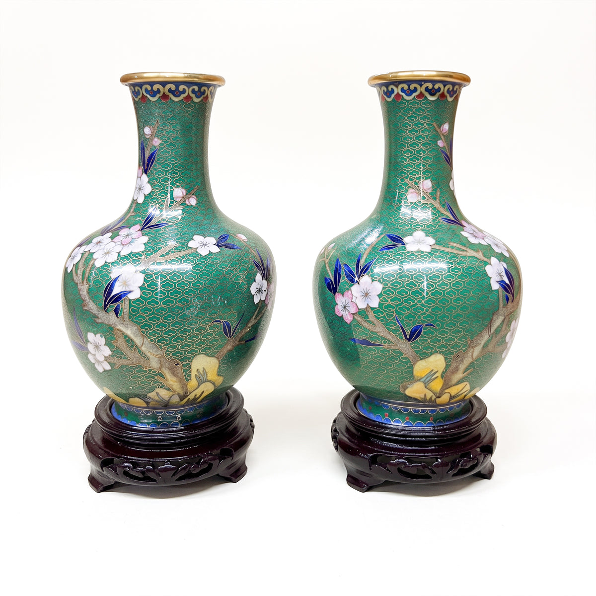 Symmetrical pair of mid-20th century Chinese cloisonné floral vases with brass wire filigree, blooming cherry blossom motifs, and tiered dark wood pedestals isolated against a neutral background.