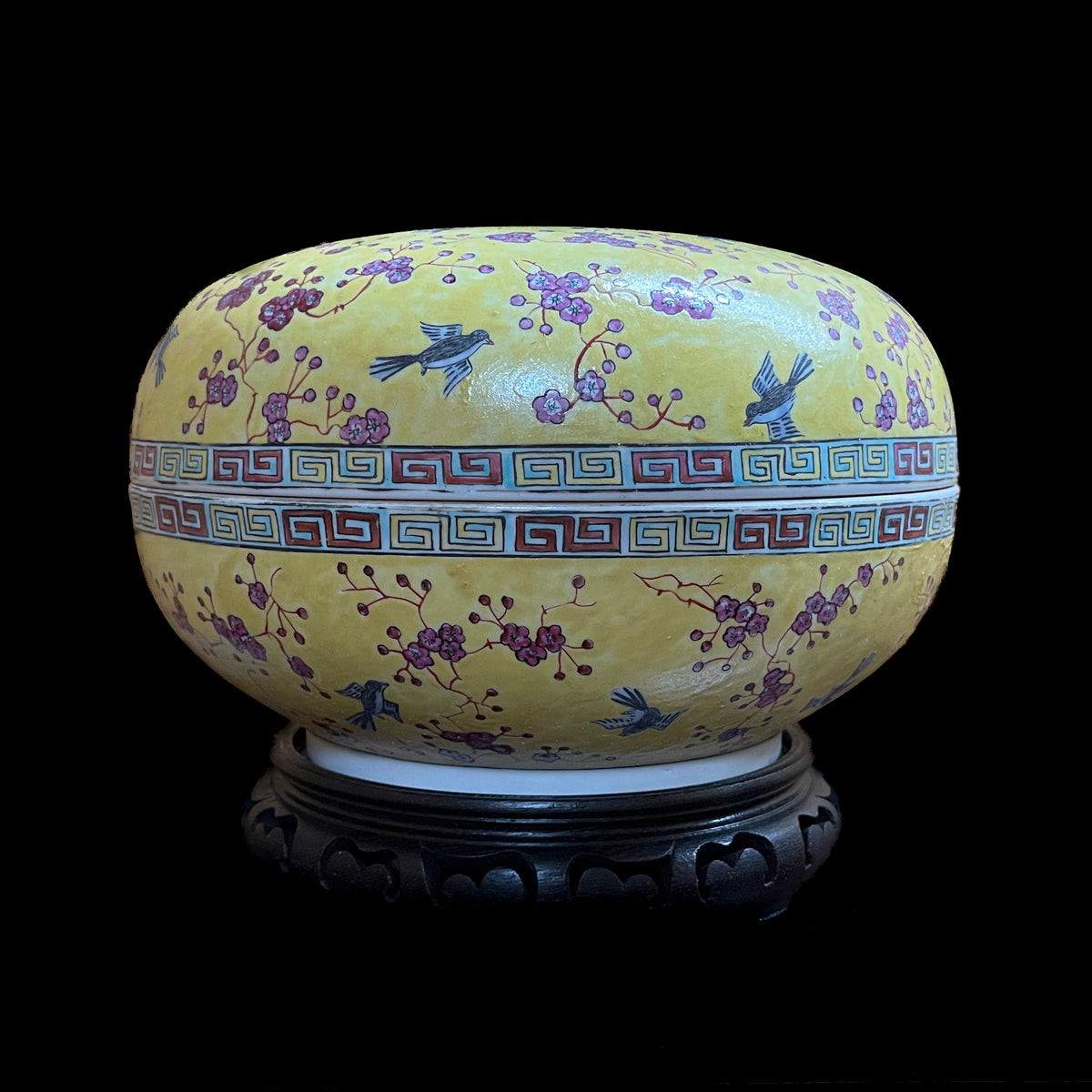 A vintage Chinese porcelain lidded sweetmeat container featuring birds and pink plum blossoms over an imperial yellow ground with a geometric key fret border on a carved wood stand.
