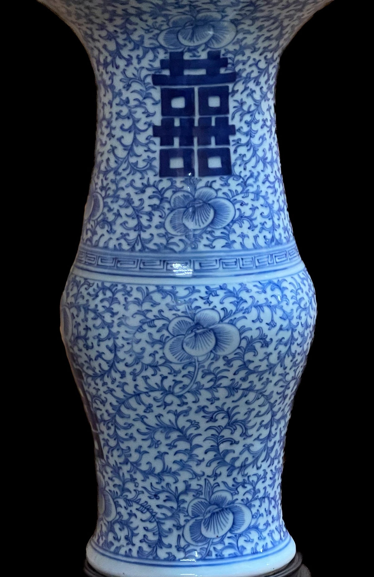 A antique vase featuring a white base with an intricate blue floral pattern. The design consists of repeating vine-like scrolls and stylized blossoms covering the entire surface of the vessel, which has a flared neck and base.