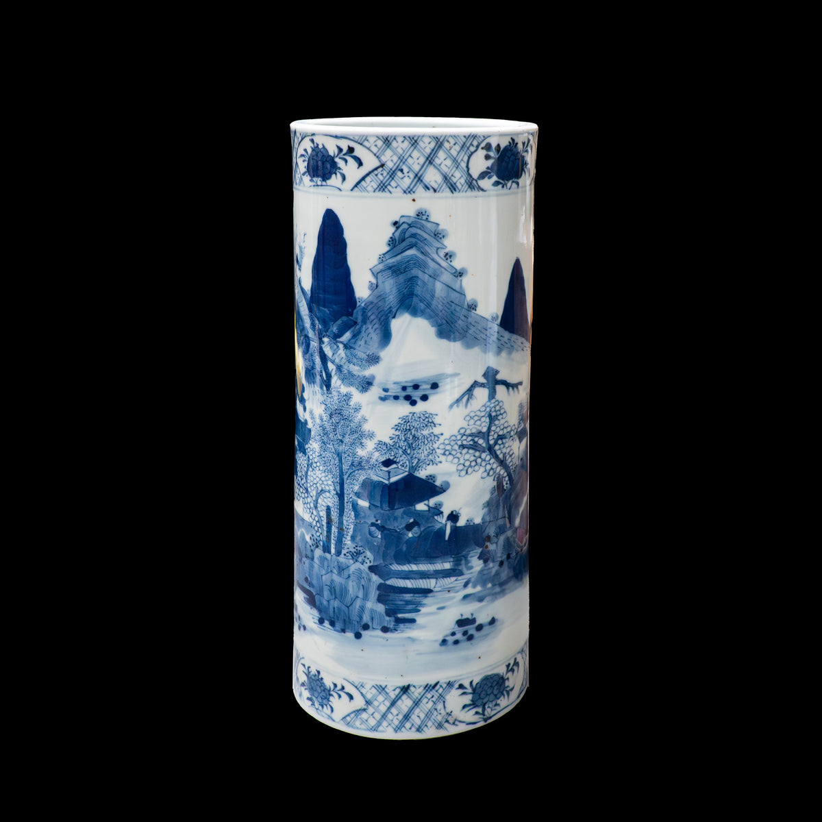 A  front view of a cylindrical 19th-century Chinese blue and white porcelain hat stand against a black background. The underglaze cobalt blue painting highlights mountain peaks rising behind a cluster of leafy pine trees and a small traditional house. Intricate cross-hatch patterned geometric borders wrap around the top and lower base.