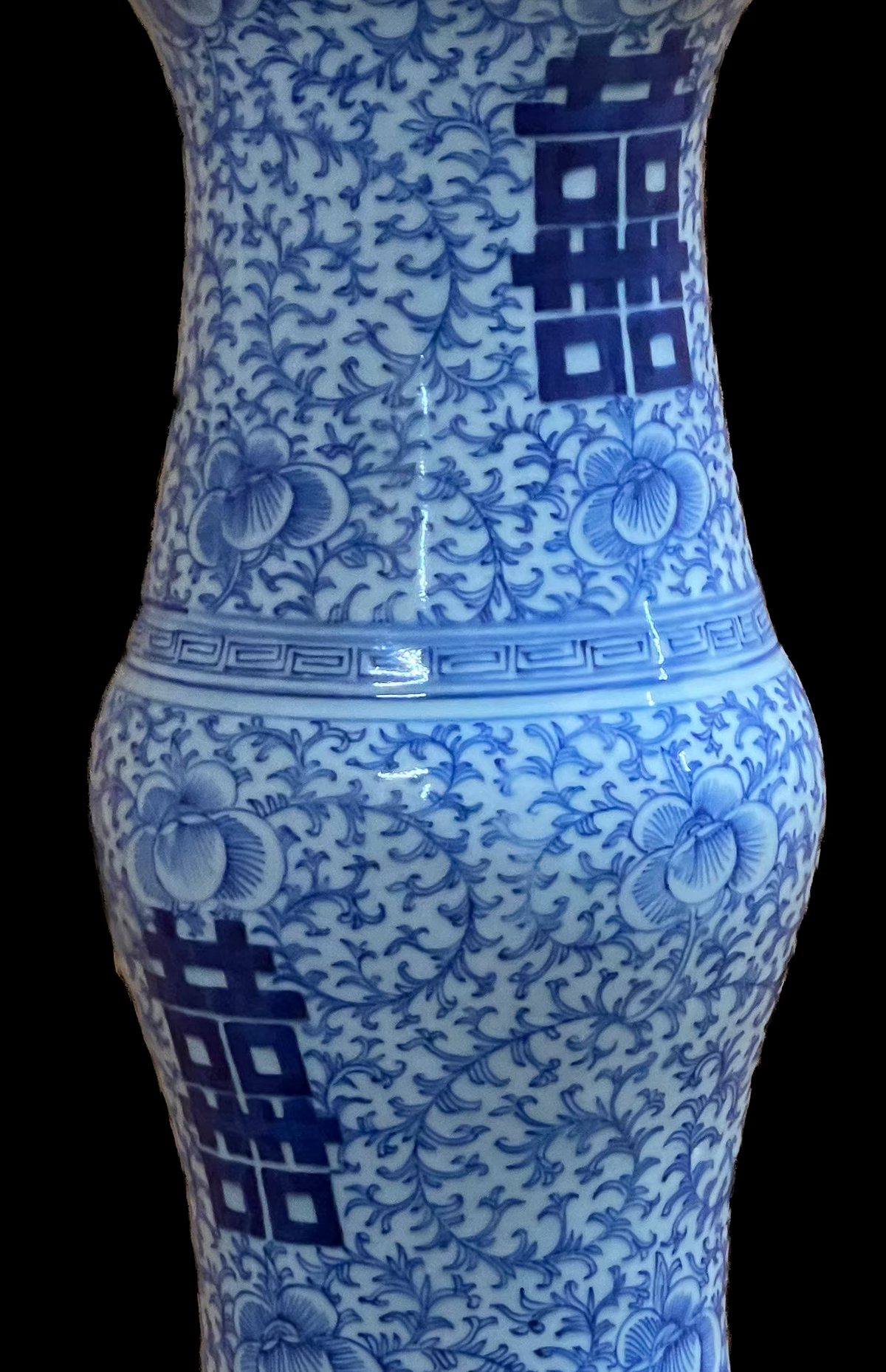 A blue and white porcelain vase close up view featuring a central double happiness character surrounded by intricate floral vine patterns. The neck of the vase flares outwards, and the base is decorated with a geometric Greek key border design.