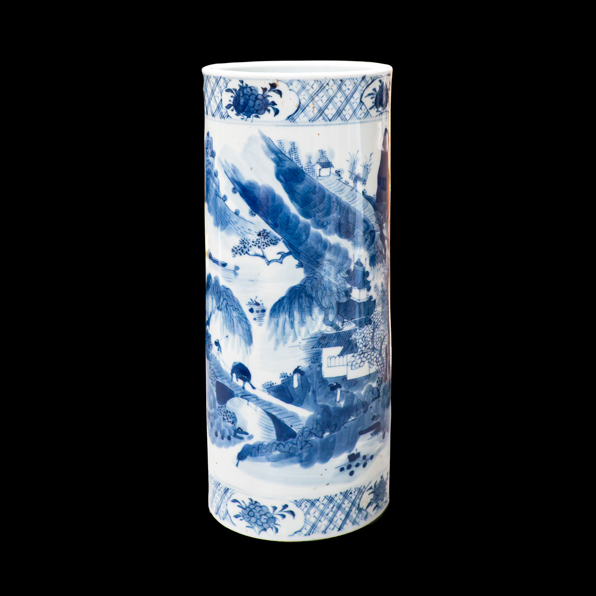 A 19th-century Chinese blue and white porcelain cylindrical hat stand against a solid black background. The hand-painted cobalt blue decoration depicts a traditional Shan Shui mountain landscape. A figure walks across a stone bridge spanning a river, leading toward a multi-story pavilion nestled in stylized hills and pine trees. Decorative cross-hatch and floral ruyi diaper bands border the top and bottom rims.