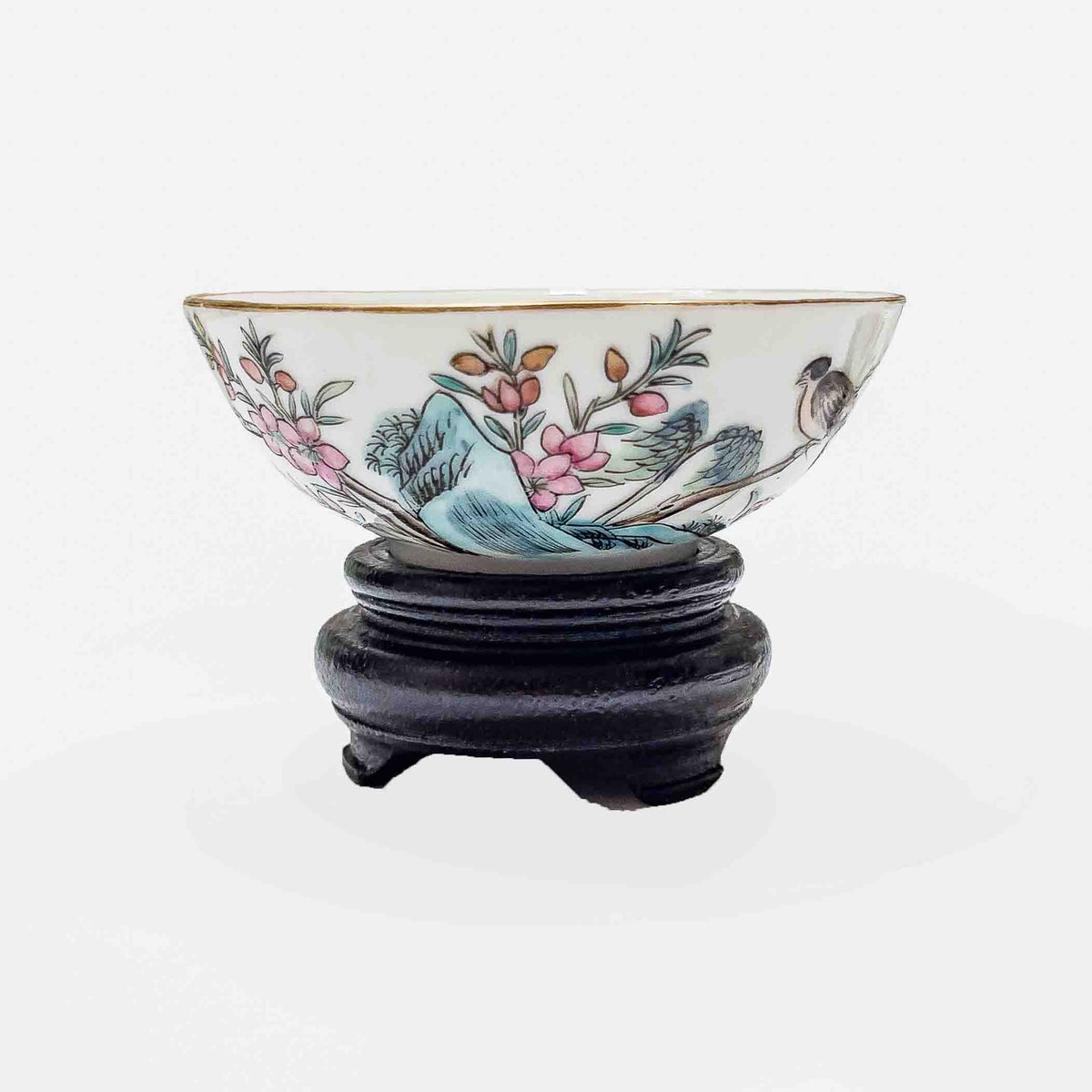 An antique Chinese famille rose porcelain 3.5 inch wine cup featuring a hand-painted songbird and pink floral blossoms with a gilded gold rim on a custom carved dark wooden base.