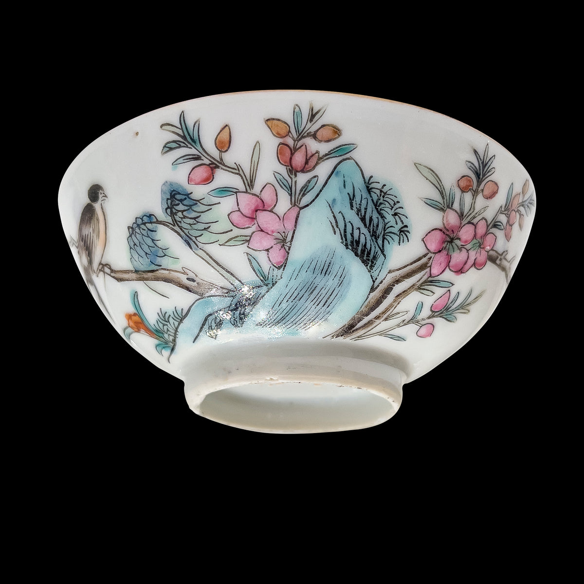 A lower angled studio shot of an antique Chinese famille rose porcelain 3.5 inch teacup showing the base foot rim and hand-enameled songbird with blue rockwork over a black ground.