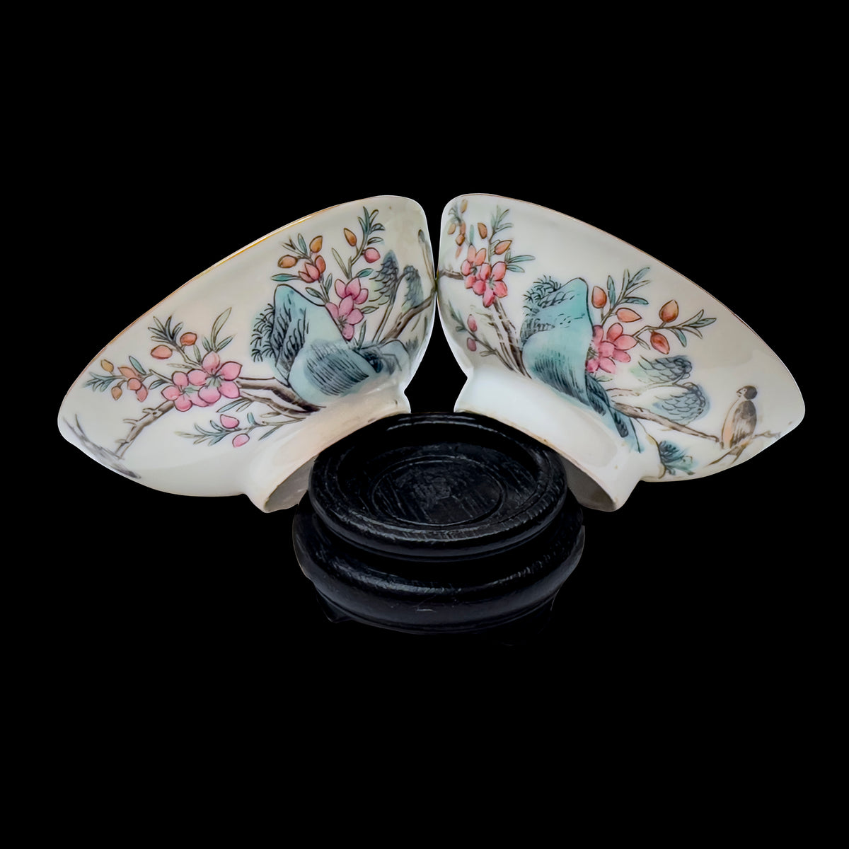 A pair of antique Chinese famille rose porcelain 3.5 inch wine teacups arranged together to show the hand-enameled bird and plum blossom motifs on a carved dark wood base over a black background.