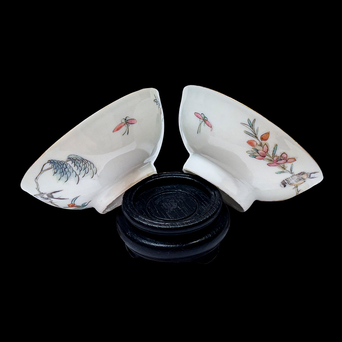 A reverse side pair shot of two antique Chinese famille rose porcelain 3.5 inch teacups showing hand-enamelled butterflies and floral branches on a carved dark wood base over a black ground