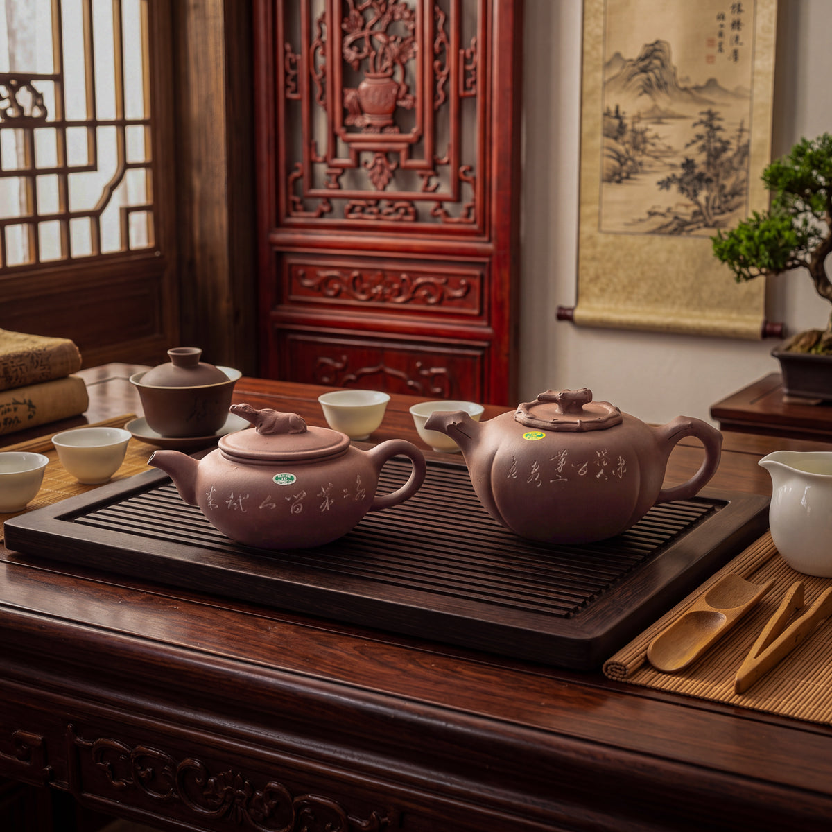Two yixing clay teapots with lids and white Chinese characters on the sides, one featuring a small animal water buffalo and the other pine branch figurine on the lid, arranged on a brown rectangular slatted wooden tea tray alongside several small white ceramic tea cups. In a beautiful traditional tea room setting.