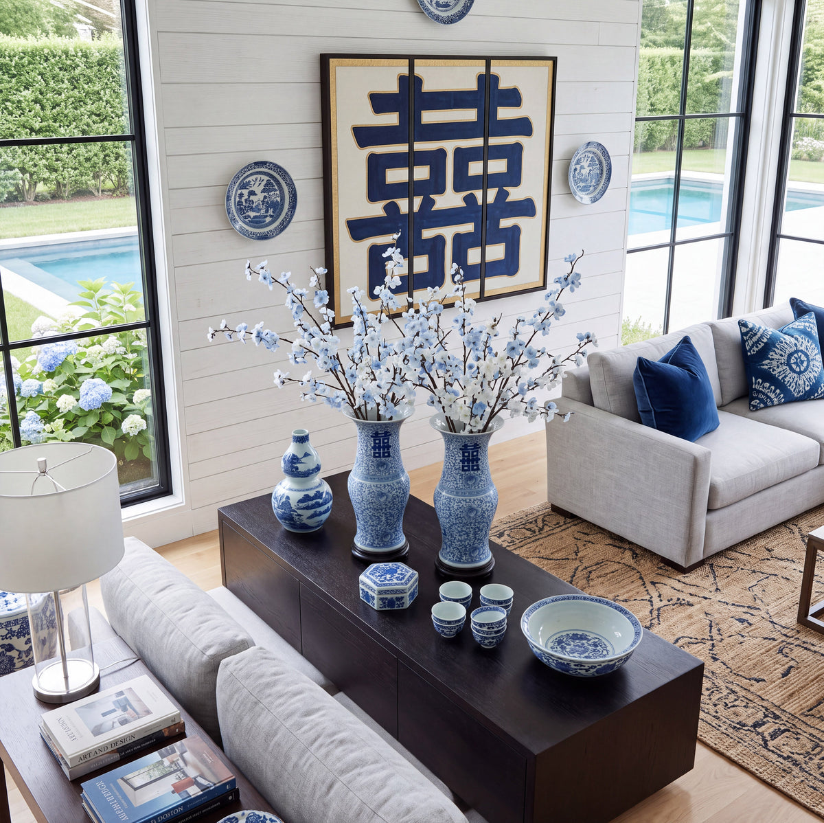 An overhead lifestyle view of a blue and white Chinese porcelain collection with double happiness vases on a dark table in a modern luxury home overlooking a backyard swimming pool.