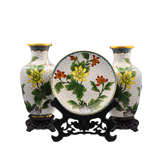 A three piece vintage Chinese cloisonne enamel garniture set featuring a pair of baluster vases and a matching decorative plate with hand-enameled yellow chrysanthemums on a white cloud ground with wooden stands.