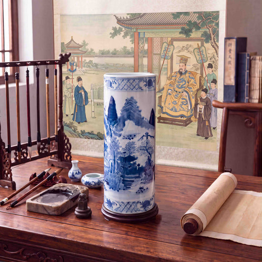 Vintage Chinese blue and white porcelain cylindrical vase featuring a painted mountain landscape scene styled on a wooden scholar desk with calligraphic brushes, inkstone, and hanging scroll.