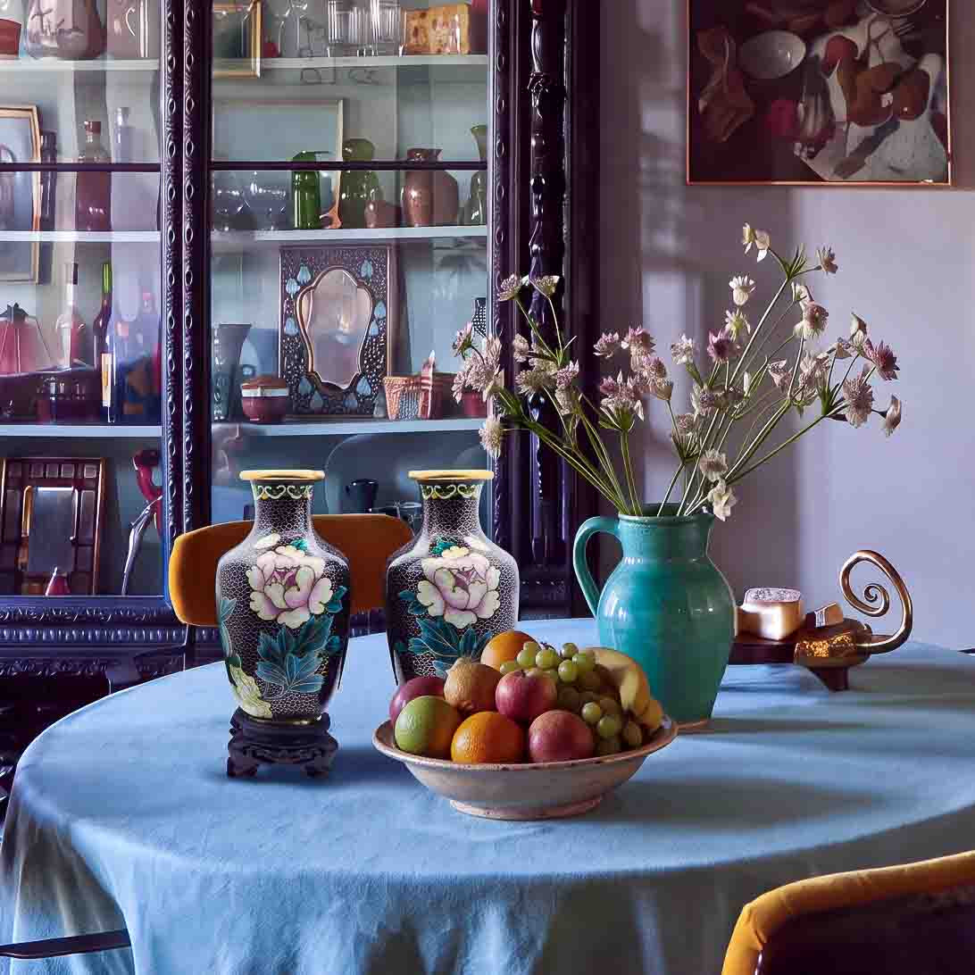 A close up photo of a pair of vintage  cloisonné enamel peony floral vases styled on a round dining table with a bowl of fresh fruit in front of an antique glass display hutch.