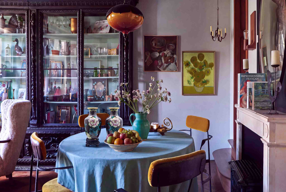 Pair of vintage cloisonné enamel vases styled on a round dining table with a blue tablecloth, fruit bowl, and turquoise vase in front of a dark antique wood glass display cabinet.