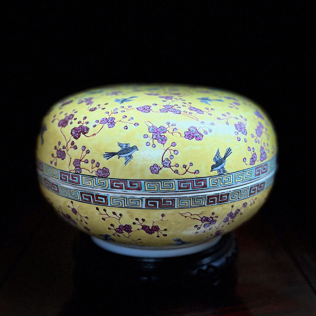 A top angled studio shot of a vintage Chinese porcelain lidded sweetmeat box showing the domed lid painted with birds and pink blossoms on an imperial yellow ground over a wood stand.